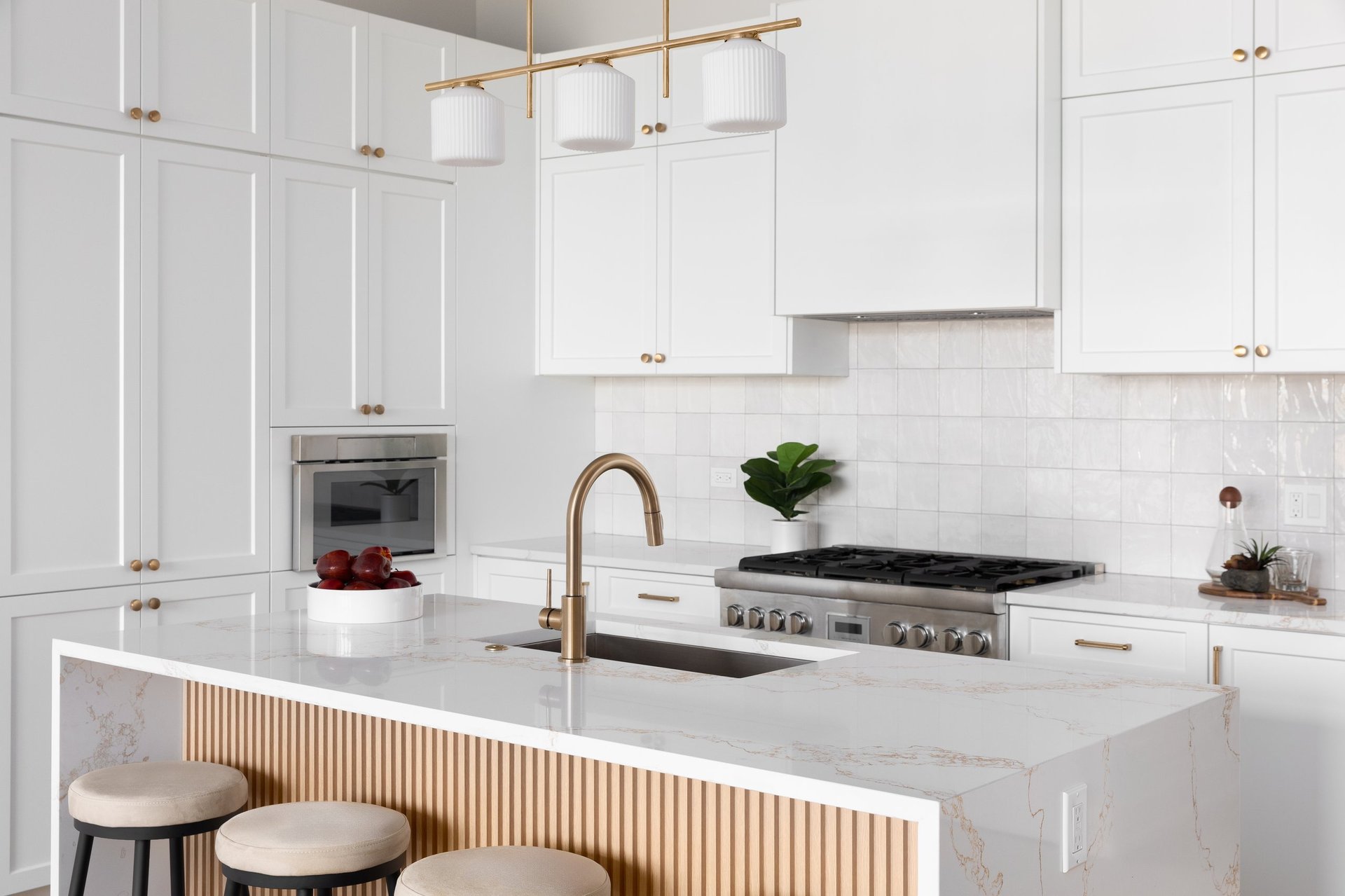 A kitchen detail with white cabinets and square tile backsplash, a waterfall marble countertop and wood slat panel front, and a gold light fixture, hardware, and faucet