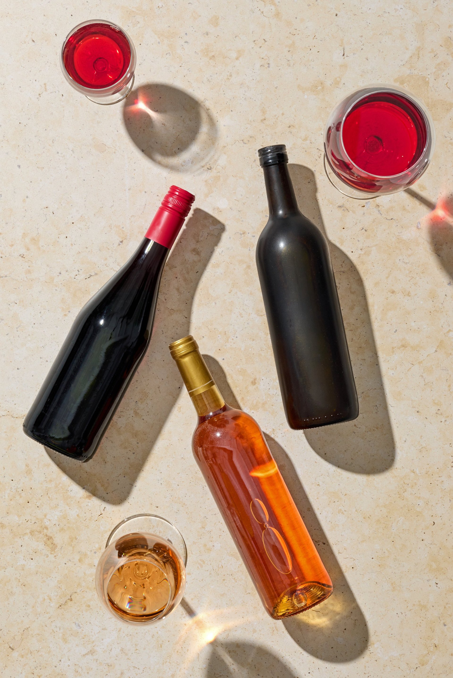 Top view of various wine bottles and wine glasses arranged on a light sunny beige background. Elegant composition with natural light and soft shadows, ideal for lifestyle, celebration and drink concepts